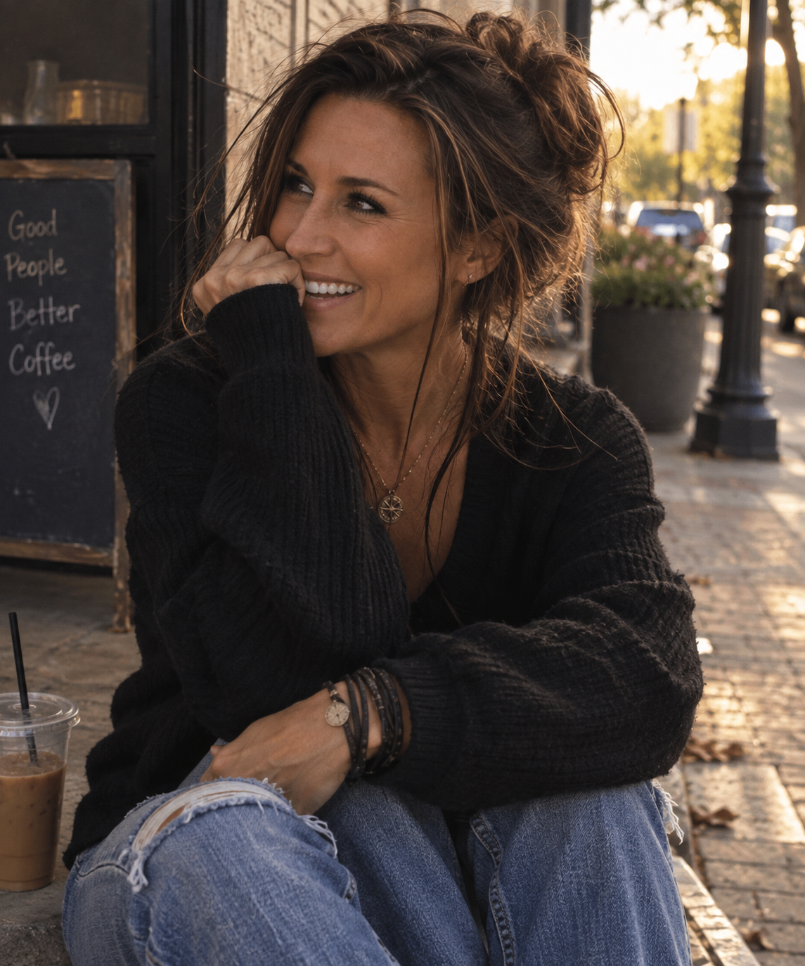 Lore Lassen laughing on a sunlit kerb outside a coffee shop, an iced coffee beside her